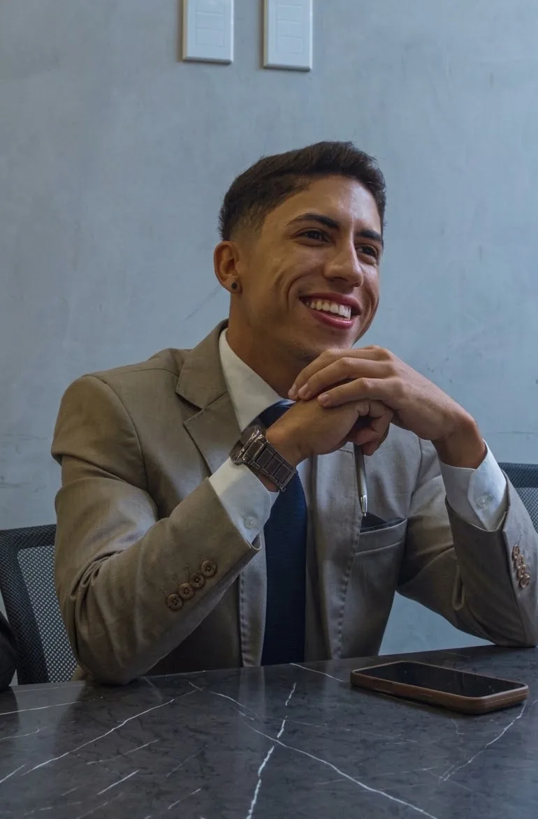 Alan Silva sentado à mesa, sorrindo, de terno bege e gravata azul